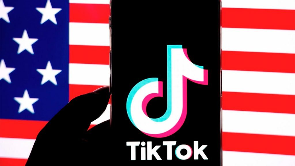 Pessoa segurando celular com logo do TikTok aberto na frente de uma imagem da bandeira dos Estados Unidos