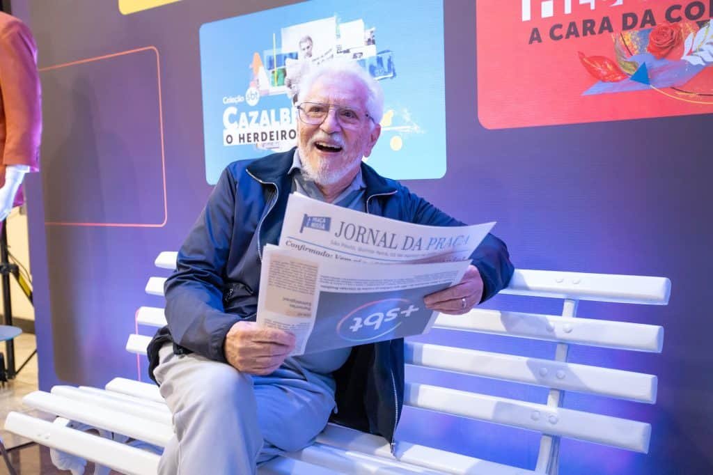 Carlos Alberto de Nóbrega sentado no banco da praça, com um jornal na mão e sorrindo